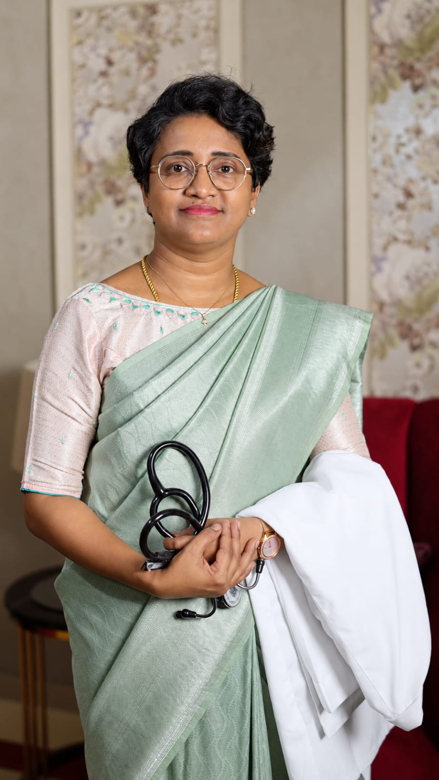 Dr. Sheela M holding a stethoscope and white coat, wearing a light green sari